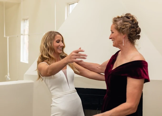 A smiling bride in a white gown opens her arms to hug her mother, who wears a burgundy velvet dress