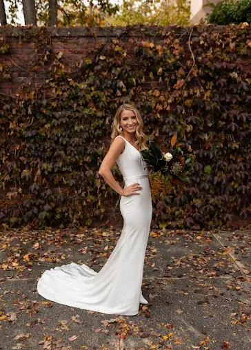 A smiling bride in a sleek gown with a train, holding a bouquet in front of an ivy wall in autumn