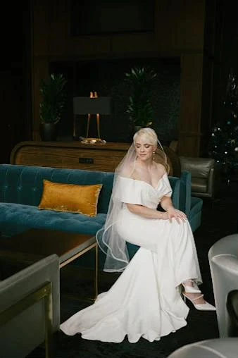 A bride in an off-the-shoulder gown with a veil, seated on a teal sofa