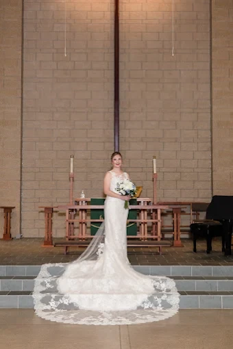 A bride in a lace gown with a long lace-edged veil and train stands in front of a church altar holding a bouquet