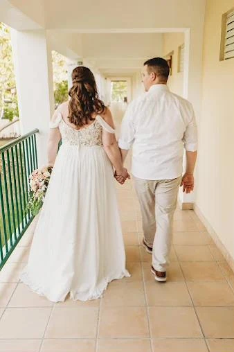 A bride and groom walk hand in hand down a sunny open-air hallway