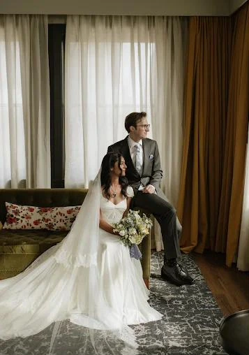 A bride with a long veil sits beside her groom by a sunlit window