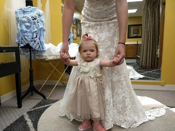 A toddler in a pink bow holds a bride’s hands during a fitting