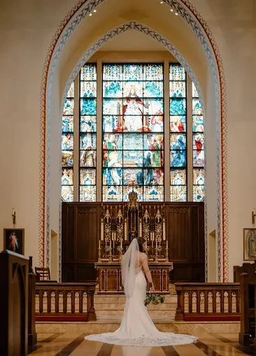 A bride in a fitted gown and long veil stands at the altar of a church beneath a stained glass window